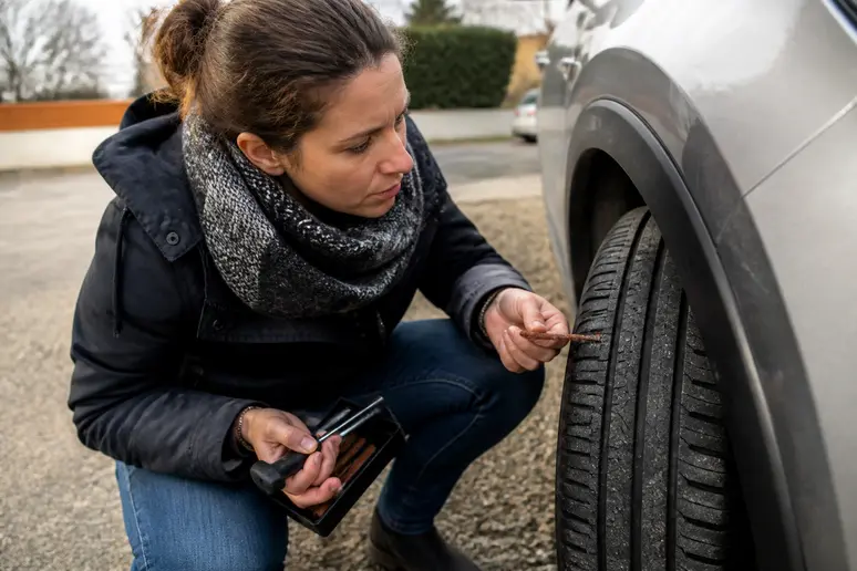 Une automobiliste française vérifie son pneu, une trousse de réparation par mèches à la main, près d'une voiture.